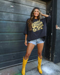 Load image into Gallery viewer, Woman wearing a black oversized sweatshirt with yellow text, denim shorts, and yellow boots standing in front of a dark metal door.