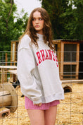 Load image into Gallery viewer, Woman wearing a gray sweatshirt with 'ARKANSAS' printed in red, standing in an outdoor setting with trees and a wooden structure. Arkansas reversible sweatshirt, razorback reversible sweater, Arkansas reversible shirt, Arkansas reversible shirt women’s, razorback store Fayetteville ar, Fayetteville boutiques, shopping near me, boutiques in the NWA mall
