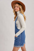 Load image into Gallery viewer, Woman wearing a denim overall dress with a white turtleneck and brown hat on a light gray background, denim romper for women, denim romper shorts, Arkansas boutiques, clothing stores Fayetteville ar