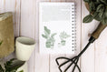 Load image into Gallery viewer, Notebook with plant illustrations on a wooden surface with gardening tools and plants.