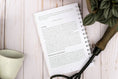 Load image into Gallery viewer, Open book with text on a wooden surface next to a plant