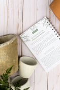 Load image into Gallery viewer, Notebook with text on a wooden surface next to a small cup and plant