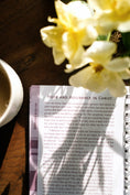 Load image into Gallery viewer, Open book with text on a wooden surface next to a cup of coffee and white flowers.