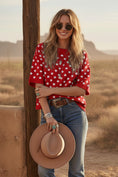 Load image into Gallery viewer, A woman smiling in the "My Hog Calling Polka Dot Sweater," a red oversized knit top with white polka dots. She is styled with a tan hat, sunglasses, and turquoise jewelry in a desert setting—a top pick for Arkansas gamedays at Whimsy Boutique.