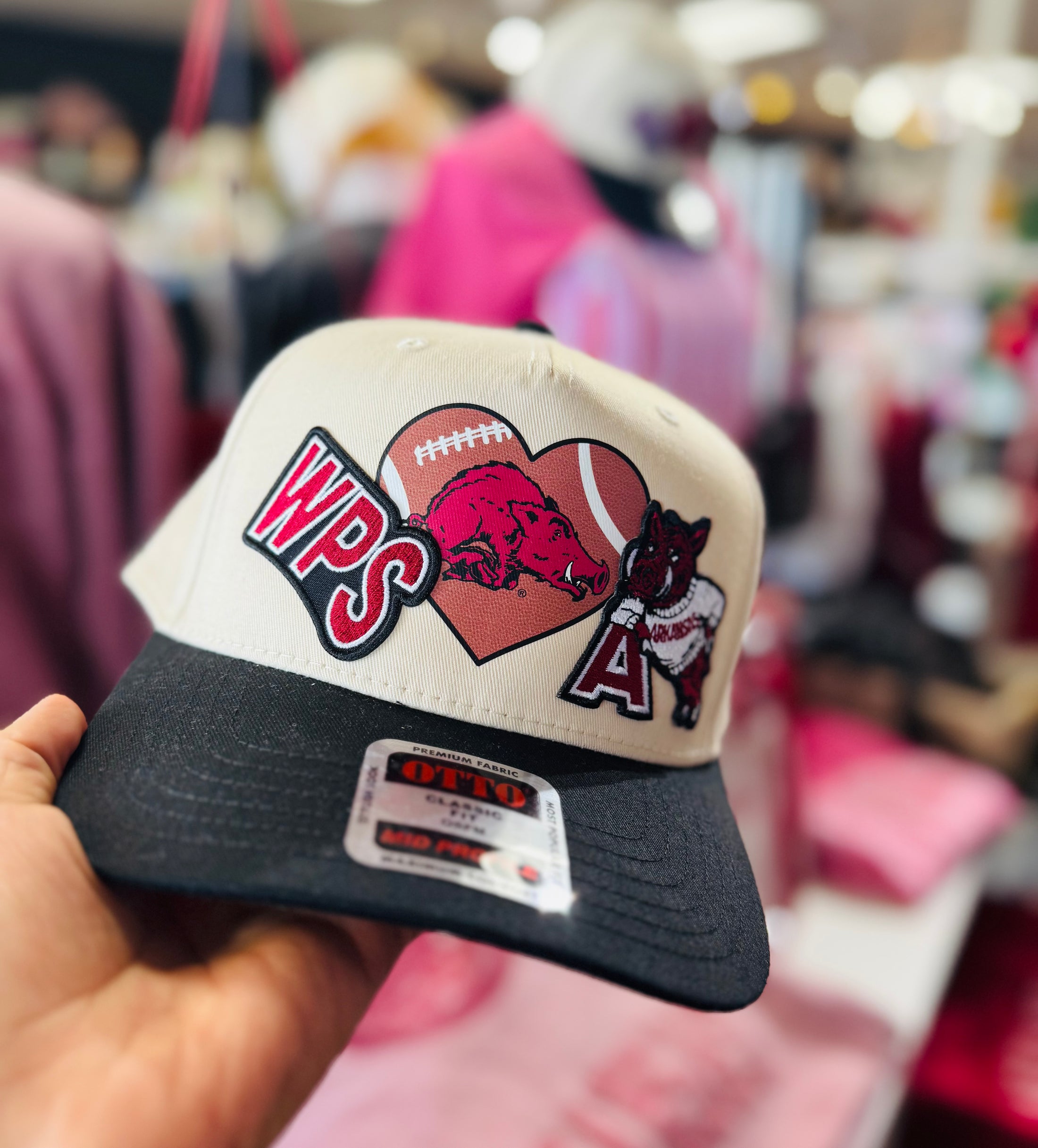 Cap with heart design and 'WPS' text held in a hand against a blurred store background, razorback hat, razorback patch hat, Arkansas Razorback trucker, Arkansas razorback trucker hat, Arkansas boutiques 