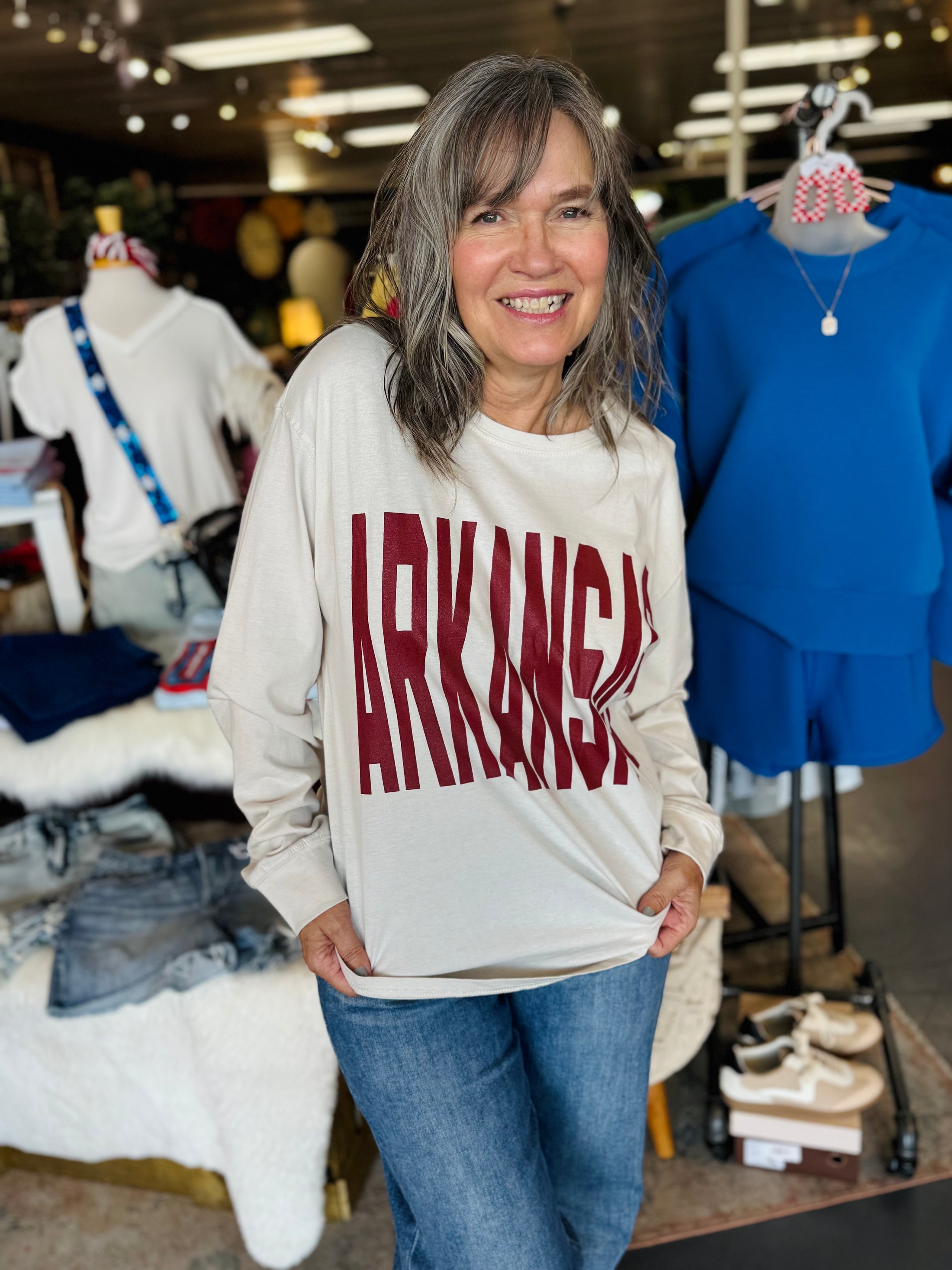 Woman wearing a 'ARKANSAS' long sleeve tee in a store setting, arkansas sweatshirt, A sweatshirt, arkansas sweatshirt women, arkansas sweatshirt near me, arkansas sweatshirt nearby, embroidered arkansas sweatshirt, university of arkansas crewneck