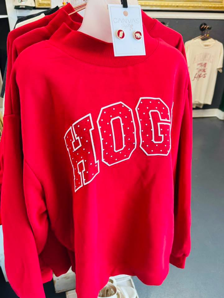 A bright red long-sleeve sweatshirt with 'HOG' printed across the chest in large white varsity-style letters outlined with small red dots. The sweatshirt is displayed on a hanger with a white retail tag attached, with other garments visible in a Fayetteville, ar boutique store Whimsy Whoo in their Razorback shop