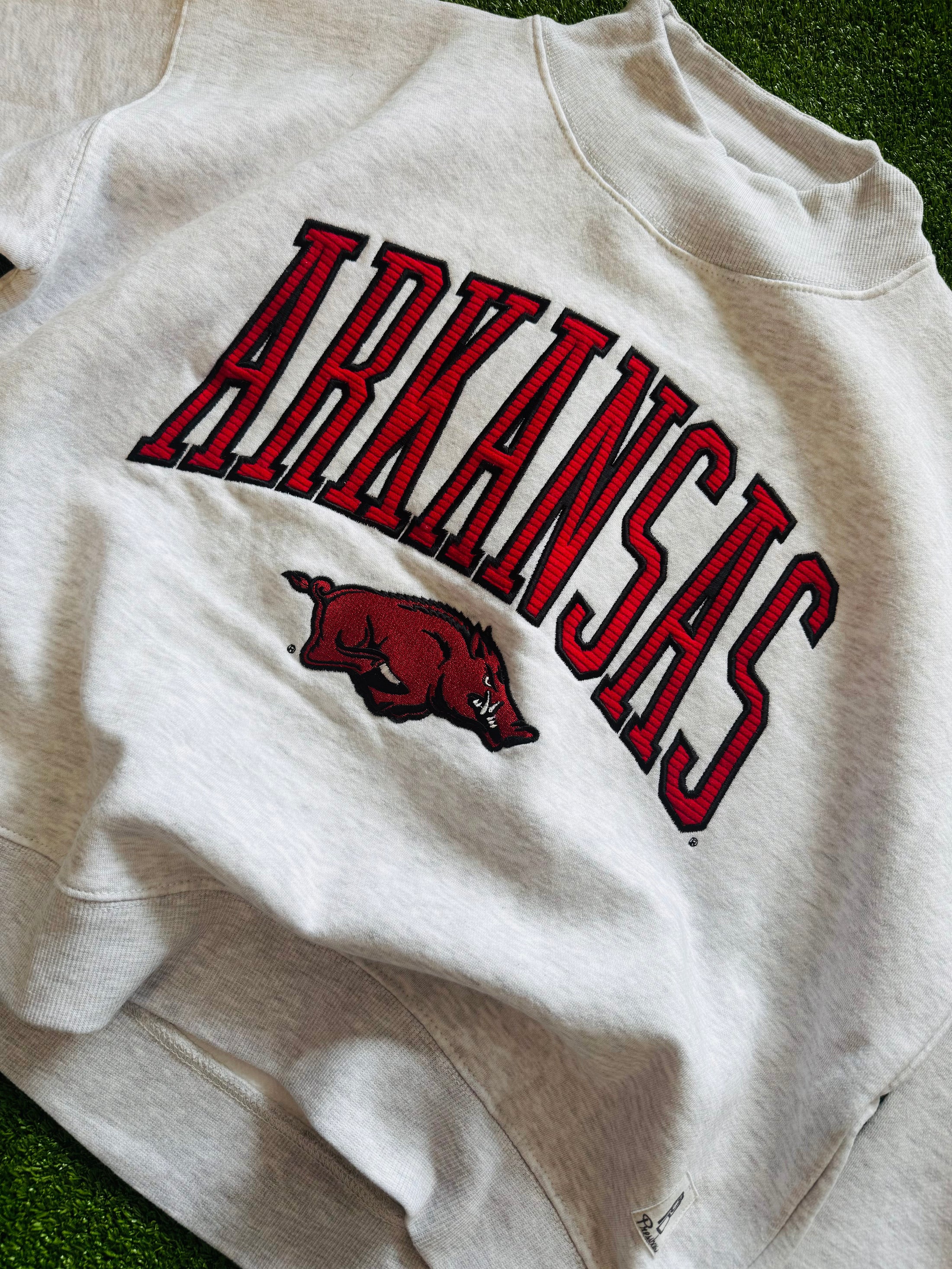 White sweatshirt with 'ARKANSAS' in red and a razorback logo on a grass background, Arkansas mock neck sweatshirt, Arkansas mock neck, arkansas razorback women’s clothing, arknasas razorback boutique clothing, university of Arkansas clothing, H&M