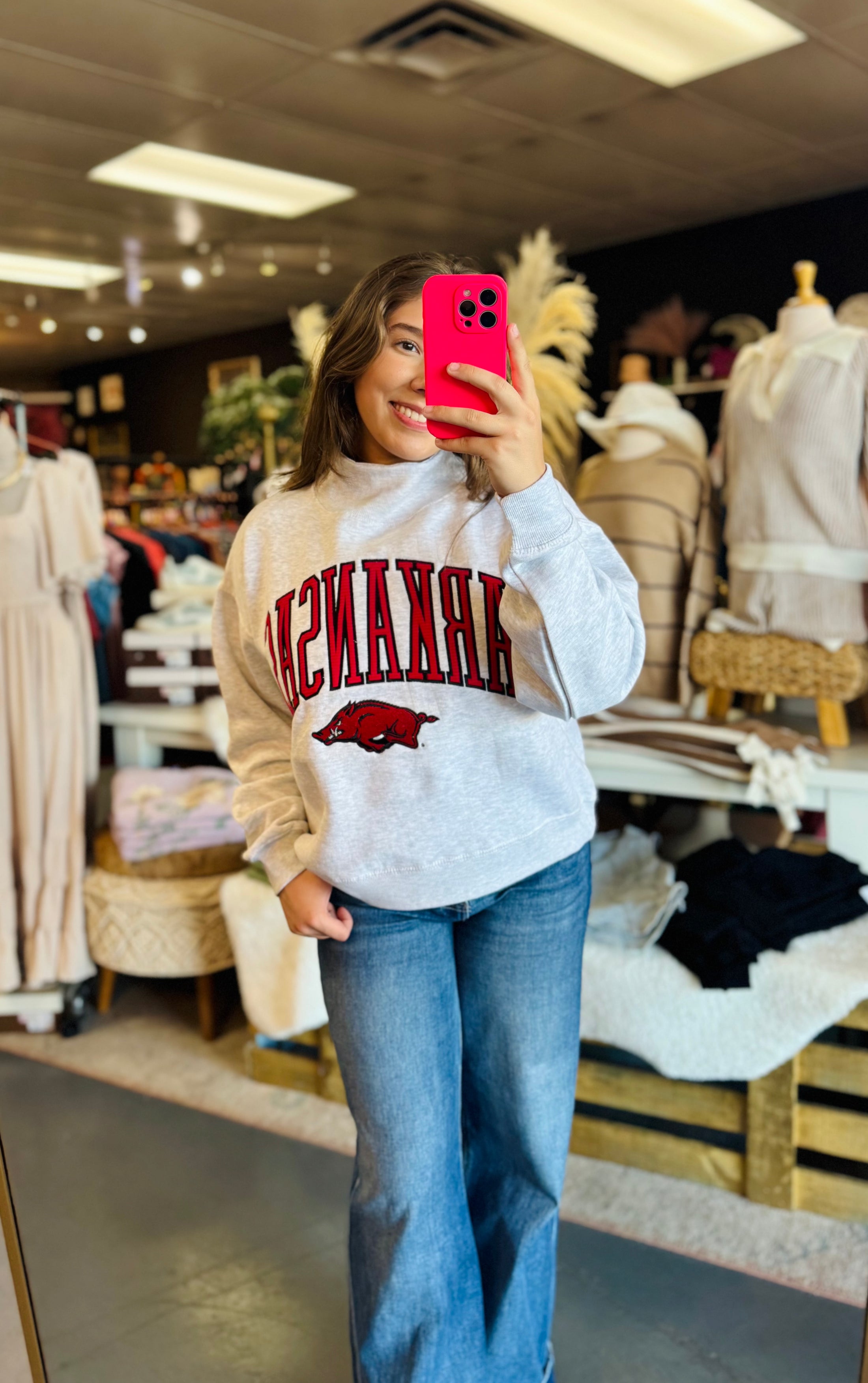Women wearing a white sweatshirt with 'University of Arkansas' text and Hogs sweatshirt, taking a mirror selfie in a Razorback store in Fayetteville ar., Arkansas sweatshirts for women near me, razorback boutique clothing near me, razorback shoppe