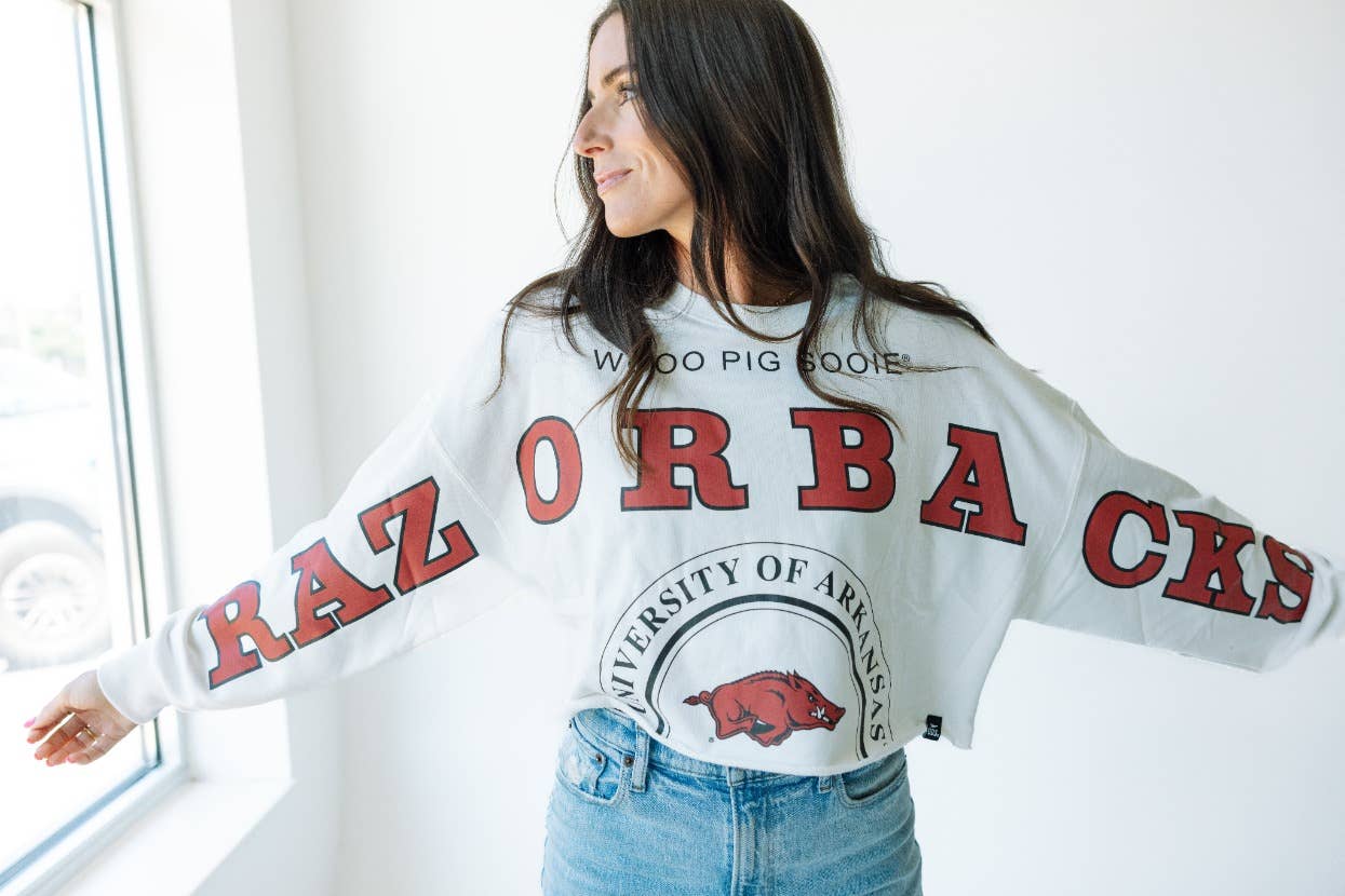 A woman wearing a white cropped long-sleeve top with 'RAZORBACK' text and the University of Arkansas at Fayetteville logo in red and black. Arkansas razorback store, cropped arkansas razorback long sleeve top, 