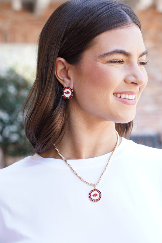 Woman wearing red circular earrings and a necklace, smiling outdoors.  Razorback jewelry, razorback jewelry near me, razorback jewelry and accessories, Arkansas razorback jewelry, razorback charms for jewelry, razorback jewelry for sale, women razorback jewelry, Arkansas razorback pendant