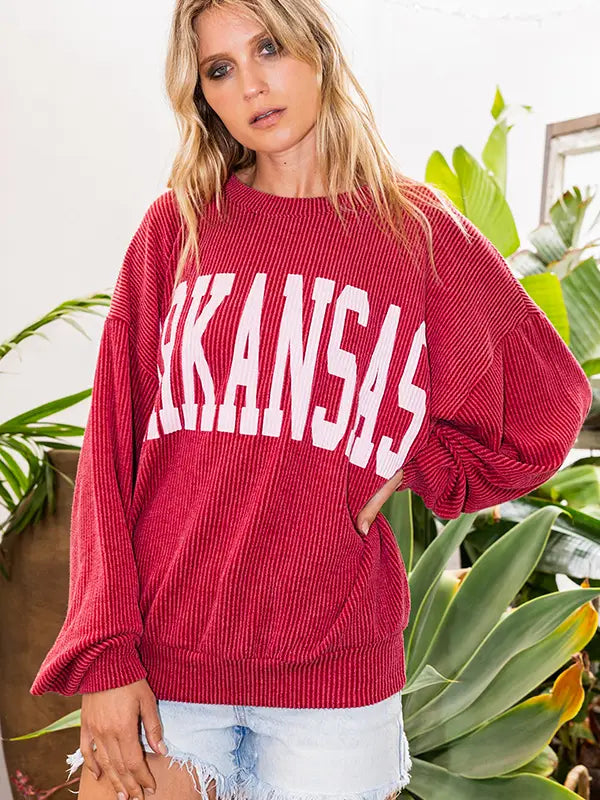 Woman wearing a red 'ARKANSAS' sweater with plants in the background, Arkansas razorback sweatshirt, Arkansas razorback sweatshirt women’s, Arkansas ribbed sweatshirt, Arkansas razorback apparel near me, Arkansas razorback store near me