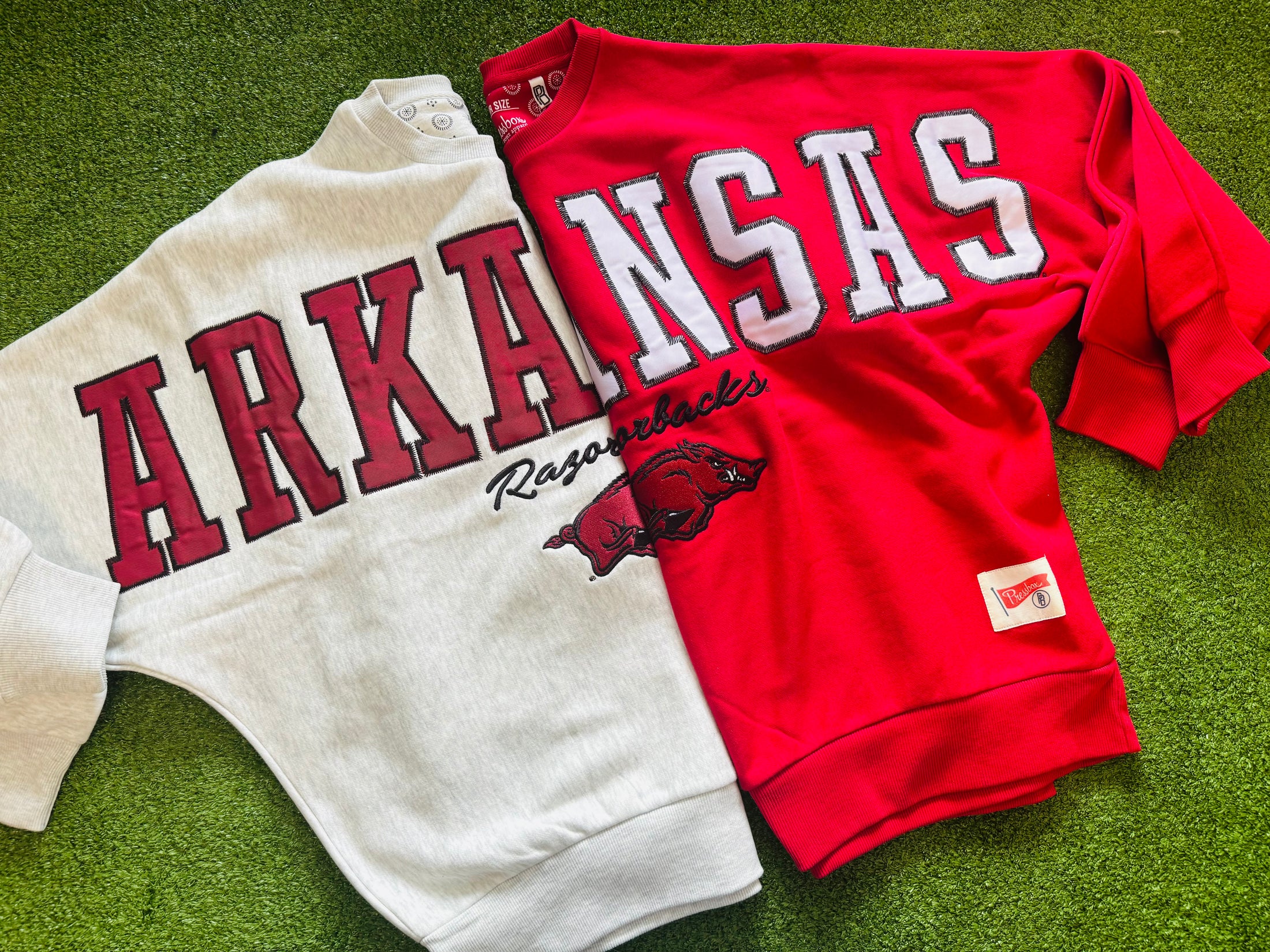Two sweatshirts, one white and one red, with 'ARKANSAS Razorbacks' text on a grass background. Razorback store in Fayetteville arkansas, arkansas sweatshirts women, university of arkansas crewneck, arkansas razorback store, store near me, Fayetteville ar stores, Arkansas razorback store