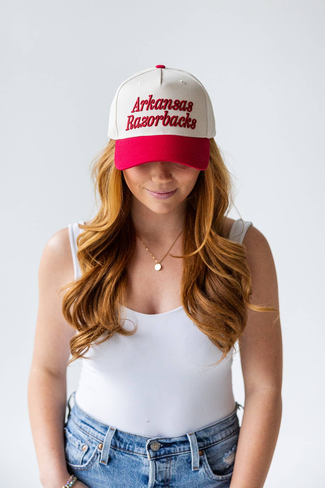Woman wearing a red and white 'Arkansas Razorbacks' cap on a light gray background, Arkansas razorback hat, razorback trucker hat, Arkansas hat, Arkansas trucker hat, Fayetteville ar boutique, Fayetteville boutique razorback clothing, razorback store