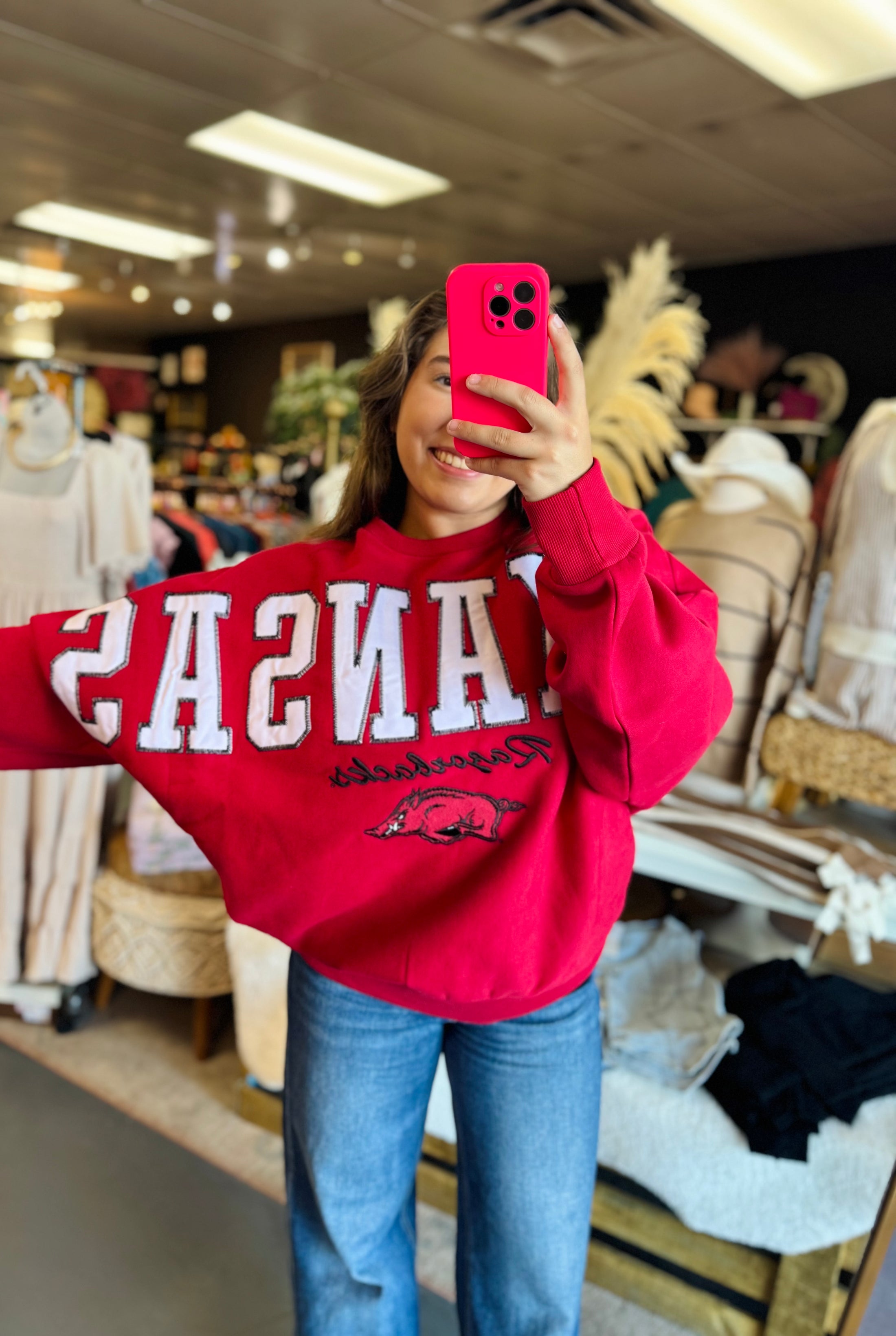 Woman wearing a red 'University of Arkansas' sweatshirt in a store. red women’s arkansas razorback sweatshirt, women’s sweatshirt for a razorback game, Arkansas sweatshirt nearby