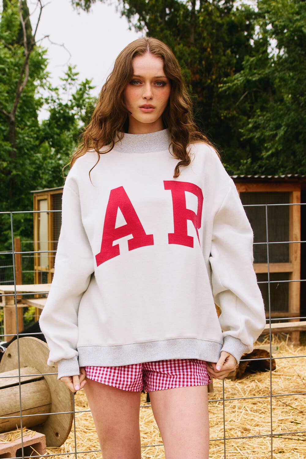 Woman wearing a white sweatshirt with red letters and checkered shorts outdoors. Arkansas reversible sweatshirt, razorback reversible sweater, Arkansas reversible shirt, Arkansas reversible shirt women’s, razorback store Fayetteville ar, Fayetteville boutiques, shopping near me, boutiques in the NWA mall