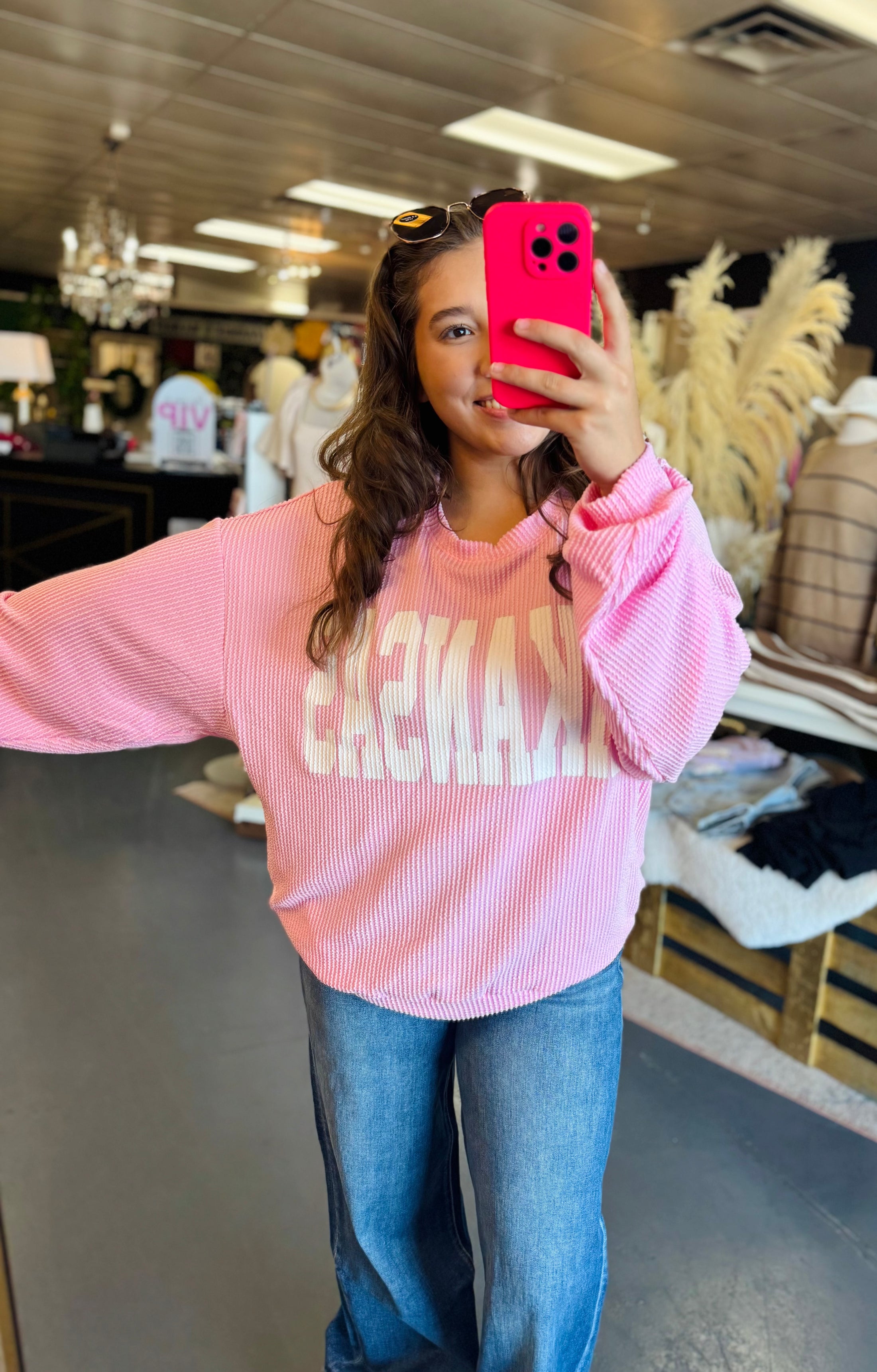 Woman wearing a pink sweatshirt with 'ARKANSAS' text, taking a mirror selfie in a clothing boutique Fayetteville ar, arkansas sweatshirt, pink arkansas sweatshirt, ribbed arkansas sweatshirt, pink ribbed arkansas sweatshirt