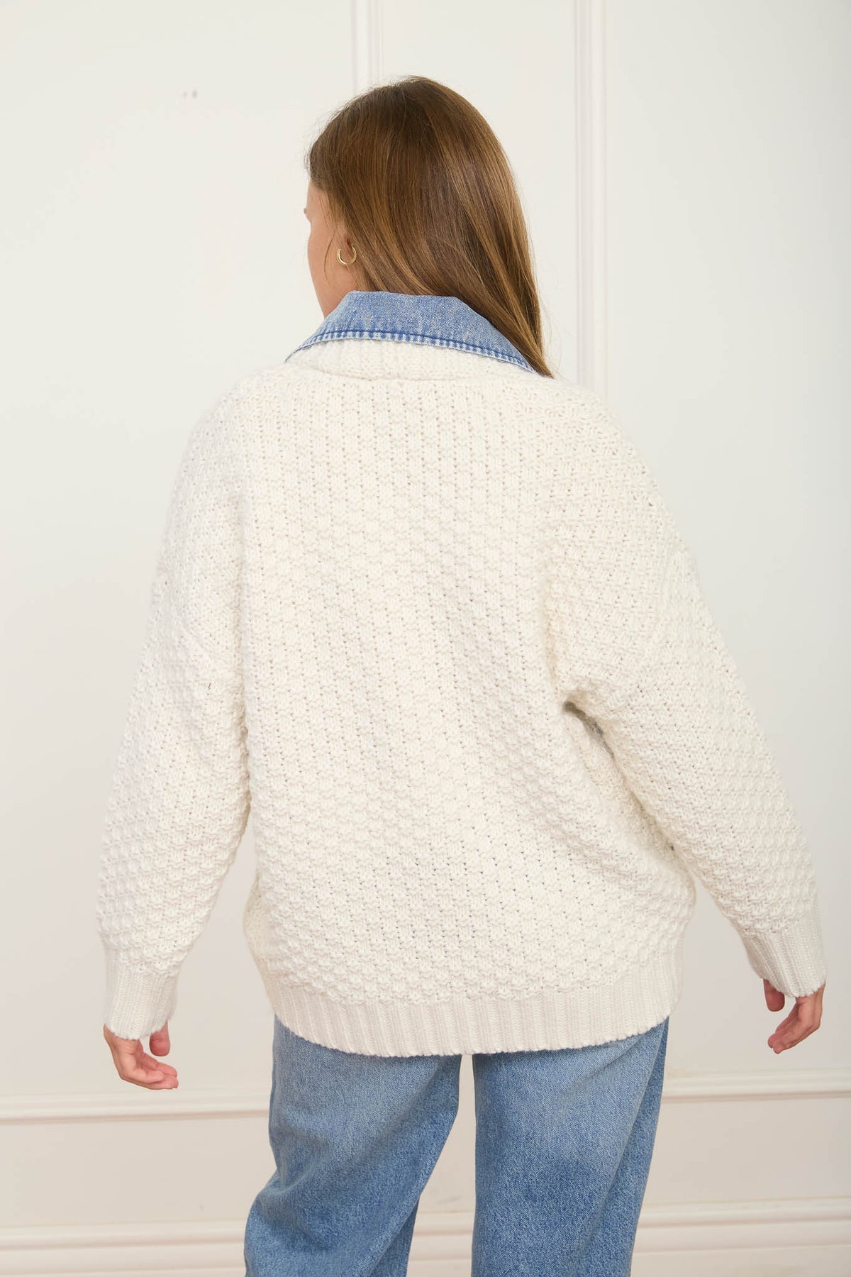 Woman wearing a white textured sweater and blue jeans in a Fayetteville Arkansas boutique store. Full-length view of the button-down long sleeve mixed media cable knit jacket with denim trim, a trending national outerwear style for women.