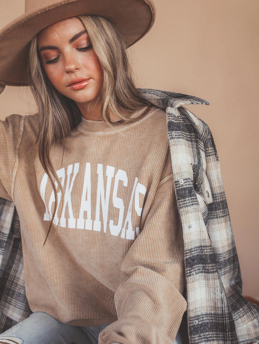 A woman wearing a beige corduroy sweatshirt with 'ARKANSAS' printed in white across the chest, styled with a brown wide-brim hat, a black-and-white plaid flannel shirt draped over her shoulder, and light blue jeans from Whimsy Whoo Boutique Store in Fayetteville, AR. 