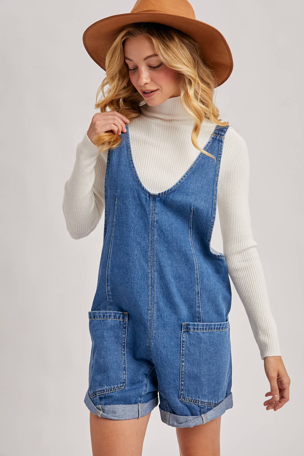 Woman wearing a denim overall romper with a white turtleneck and brown hat on a light gray background, denim romper, denim romper for women, Arkansas boutiques 