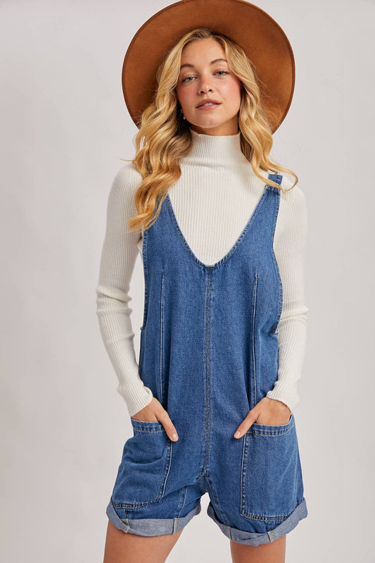 Woman wearing a denim romper with a white turtleneck and brown hat on a light gray background, denim romper shorts, denim romper near me, Fayetteville ar boutique, Arkansas boutiques 