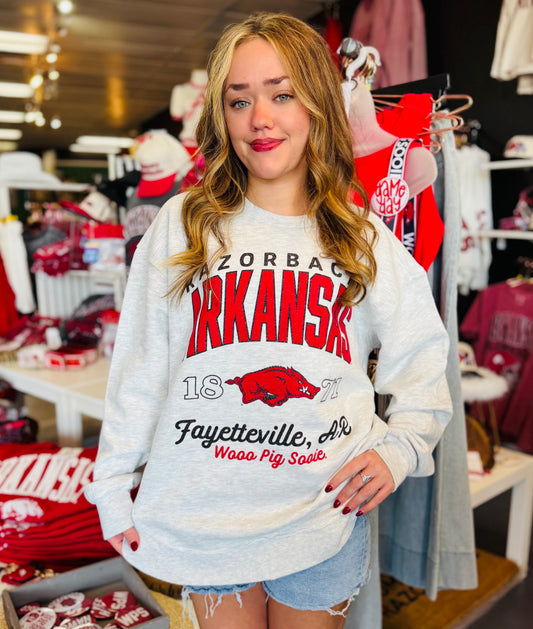 Woman wearing a gray sweatshirt with Arkansas-themed design in a store setting, gray Razorback sweatshirt, Pressbox Arkansas sweatshirt, gray Arkansas sweatshirt, razorback shirts, Arkansas shirts, AR Razorback apparel, Razorback stores Fayetteville ar shopping