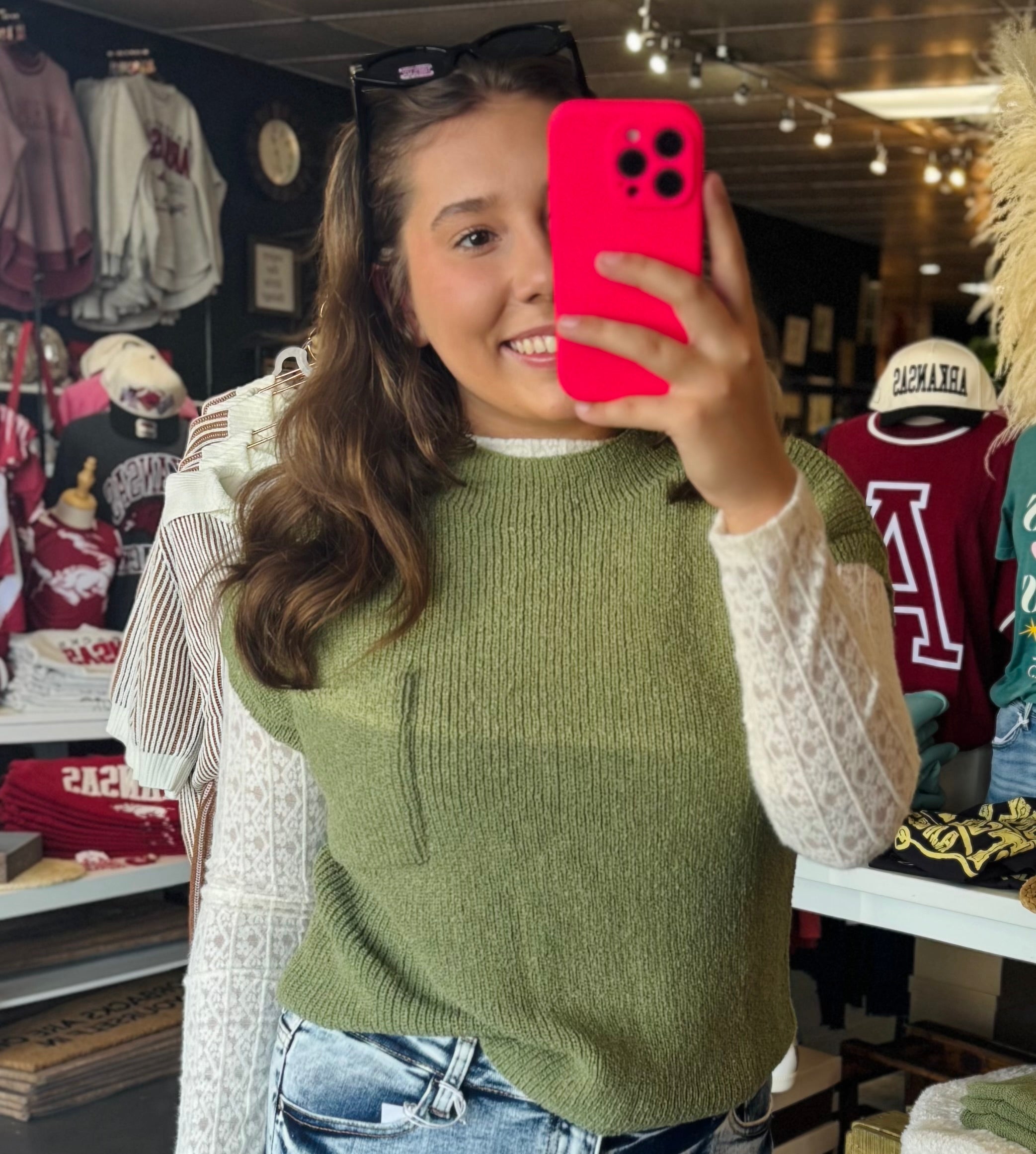 Women  taking a selfie in a Fayetteville ar boutique store wearing a green sweater vest and holding a pink phone case. Green sweater, green short sleeve sweater, blu pepper sweater, boutiques near me, boutiques Fayetteville ar