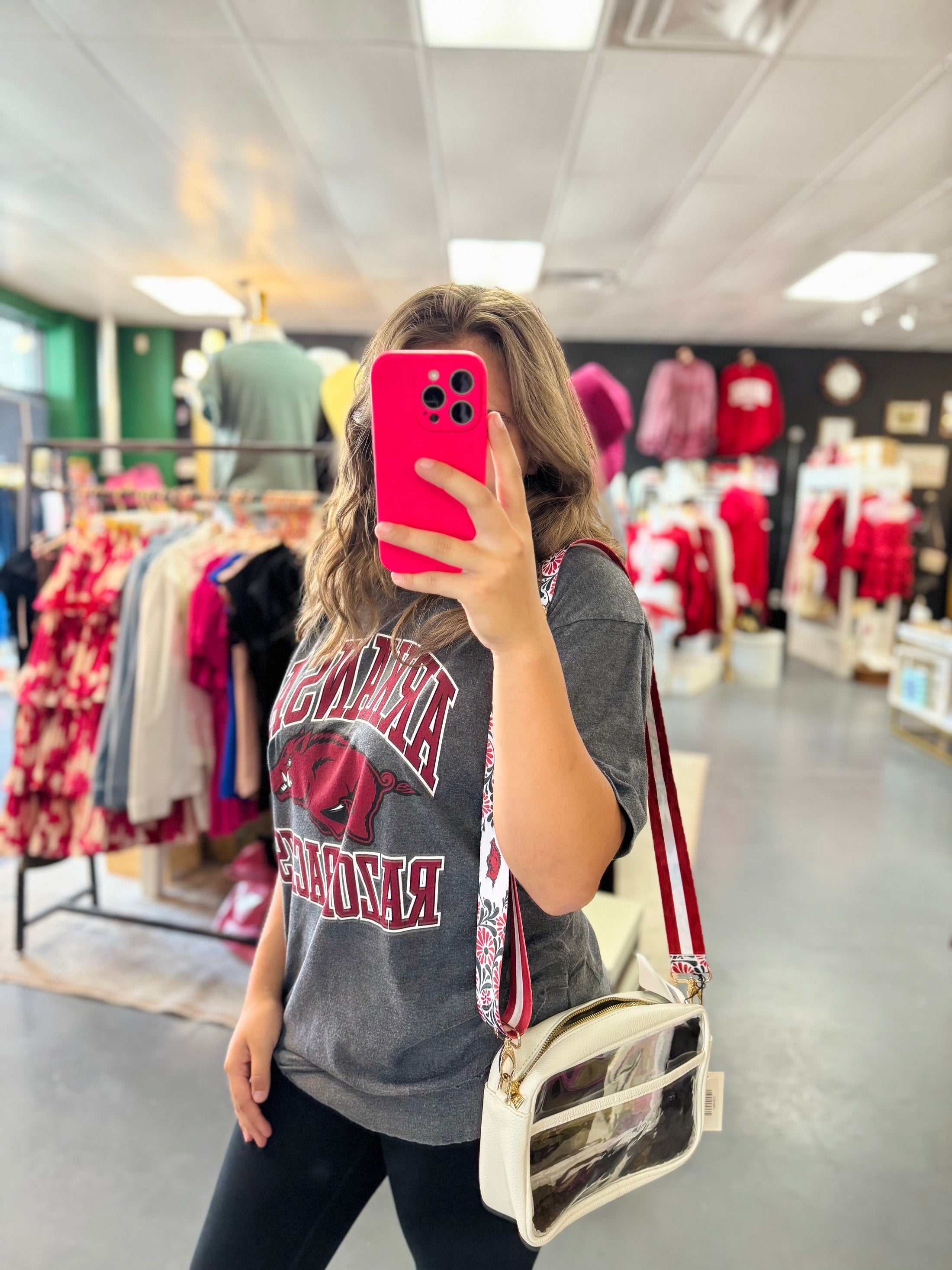 Woman taking a mirror selfie in a razorback clothing store wearing a gray 'arkansas' shirt and holding a pink phone.  grey Arkansas razorback tee, Arkansas razorback shirts, arkansas razorback boutique clothing, arkansas razorback shirts, razorback store, razorback apparel Fayetteville ar,  arkansas razorback store
