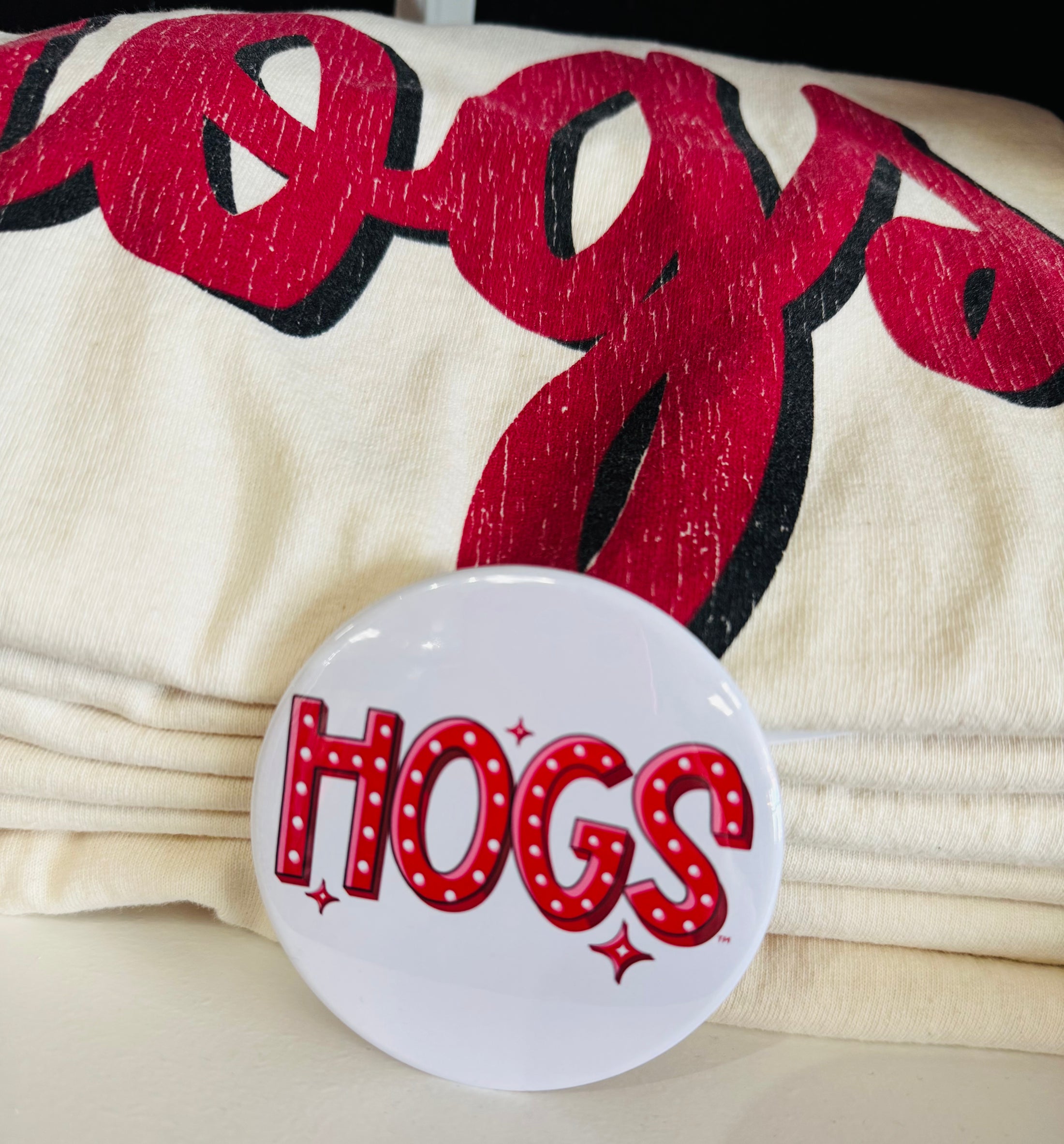 Button with 'HOGS' text on a background of red and black lettering on beige fabric. gameday buttons near me, gameday buttons sorority, gameday button pins, Arkansas buttons, art buttons, Fayetteville boutique store, shopping near me NWA mall, Arkansas gameday buttons, razorback store fayetteville
