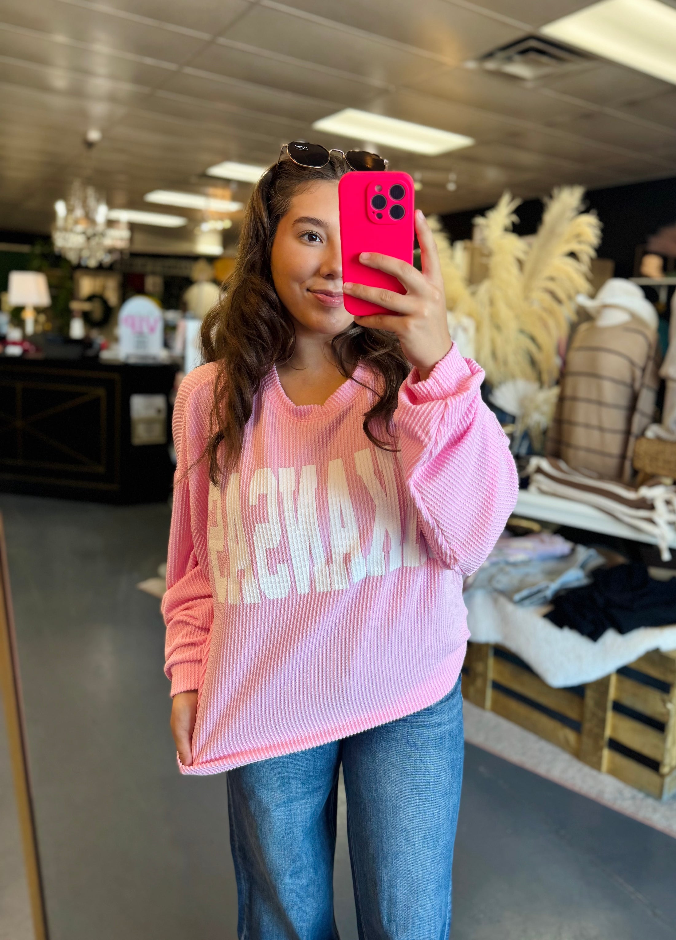 Women wearing a pink sweatshirt with text, taking a mirror selfie in a clothing store Fayetteville ar.  Pink ribbed arkansas sweatshirt, arkansas pink sweatshirt, ribbed arkansas top
