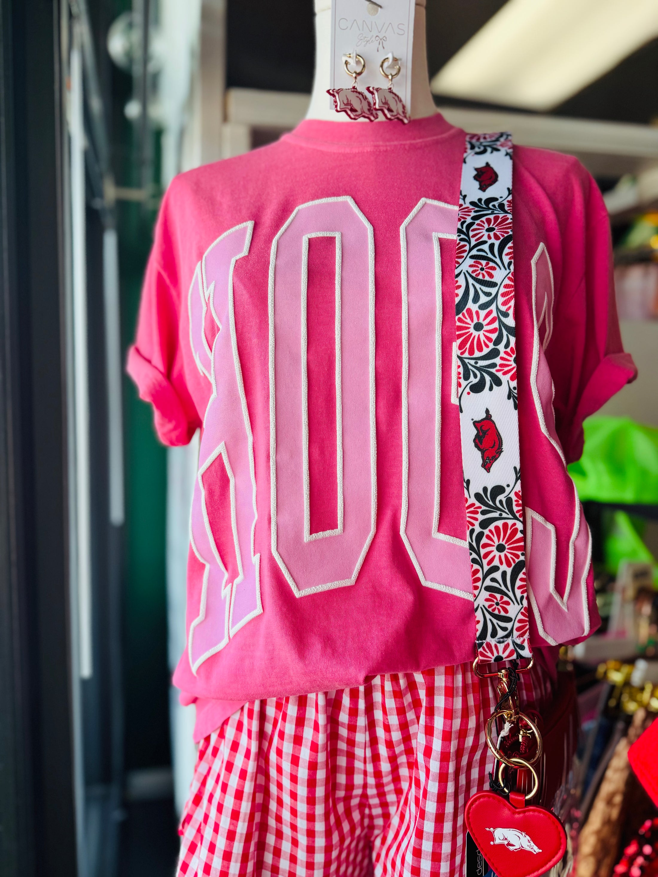 Pink shirt with large letters, red checkered pants, and a patterned strap on a mannequin, Pink hogs shirt, pink razorback shirt, women’s razorback shirt, women’s hogs shirt, razorback shirt for women, hogs shirt for women, razorback top for women, hogs top for women