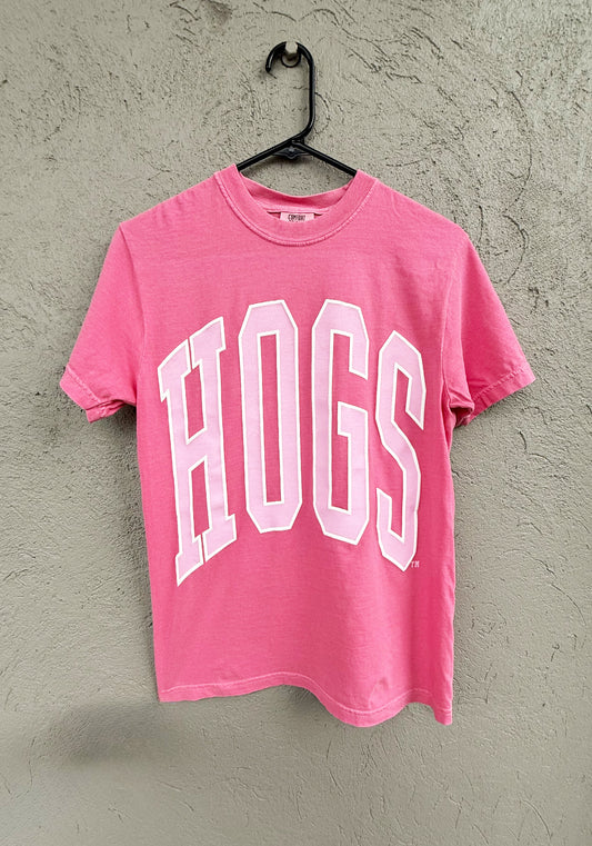 Pink t-shirt with 'HOGS' text on a hanger against a textured wall.  Pink hogs shirt, pink razorback shirt, women’s razorback shirt, women’s hogs shirt, razorback shirt for women, hogs shirt for women, razorback top for women, hogs top for women
