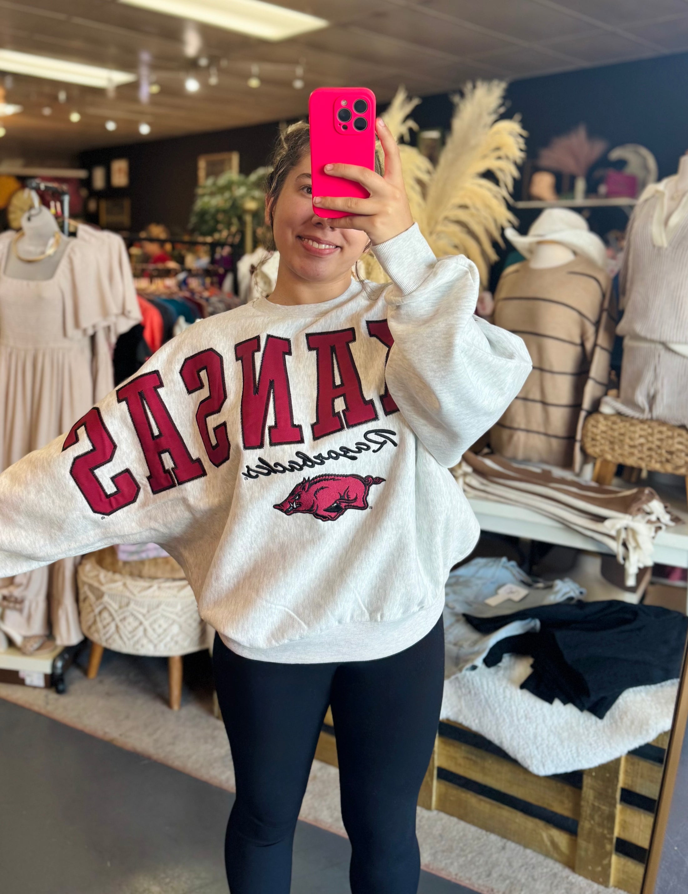Person wearing a white sweatshirt with red text and logo in a store setting, Pressbox sweatshirt Fayetteville, arkansas sweatshirts near me, razorback sweatshirt women near me, razorback shop Fayetteville ar, Arkansas clothing near me, arkansas razorback store