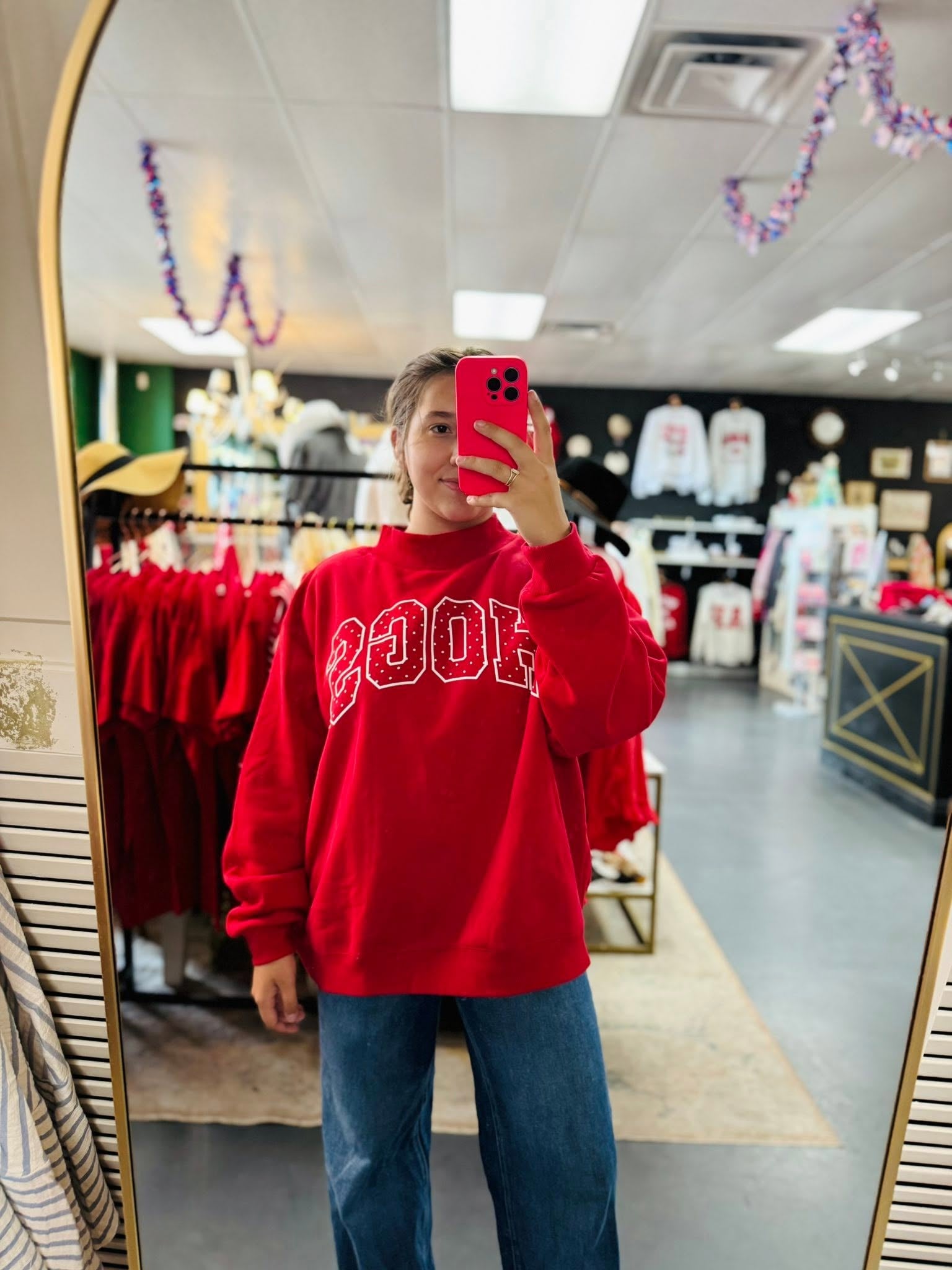 A person taking a mirror selfie in a clothing store, wearing a bright red sweatshirt with 'HOGS' printed in white across the chest and blue jeans.