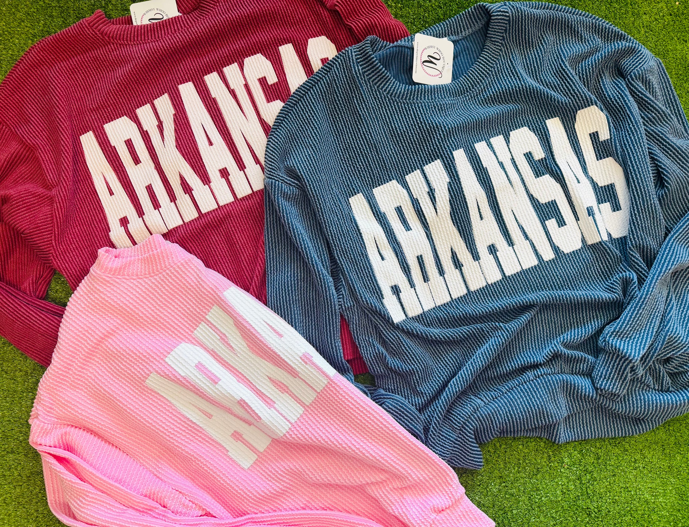 Three sweatshirts with 'ARKANSAS' printed on them, one red, one blue, and one pink, in a Fayetteville arkansas razorback store, razorback store, arkansas boutique, Razorbacks sweatshirt