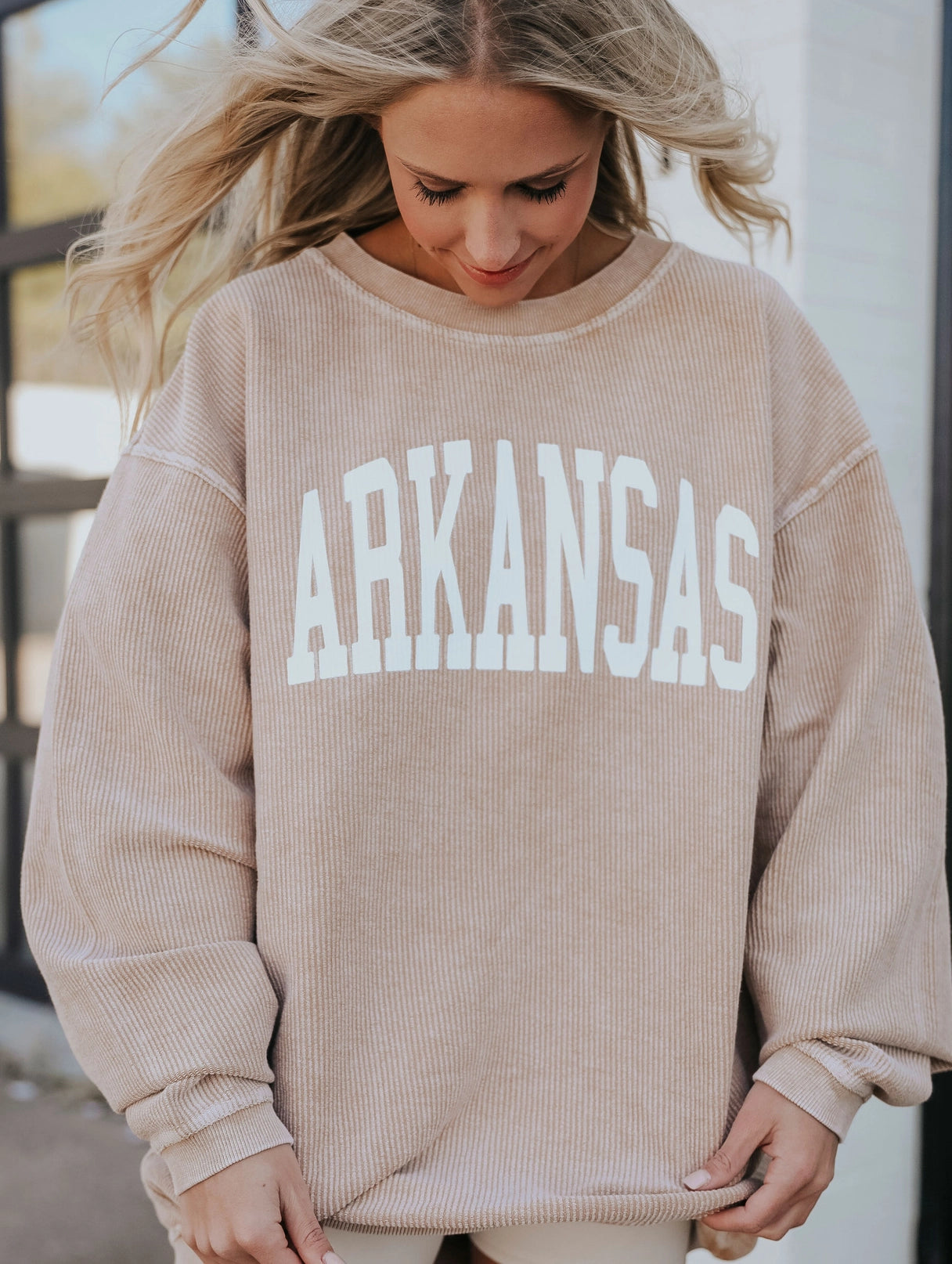 A woman wearing a beige ribbed crewneck sweatshirt with 'ARKANSAS' printed in large white block letters across the chest, from Whimsy Whoo boutique’s Razorback shop in Fayetteville.