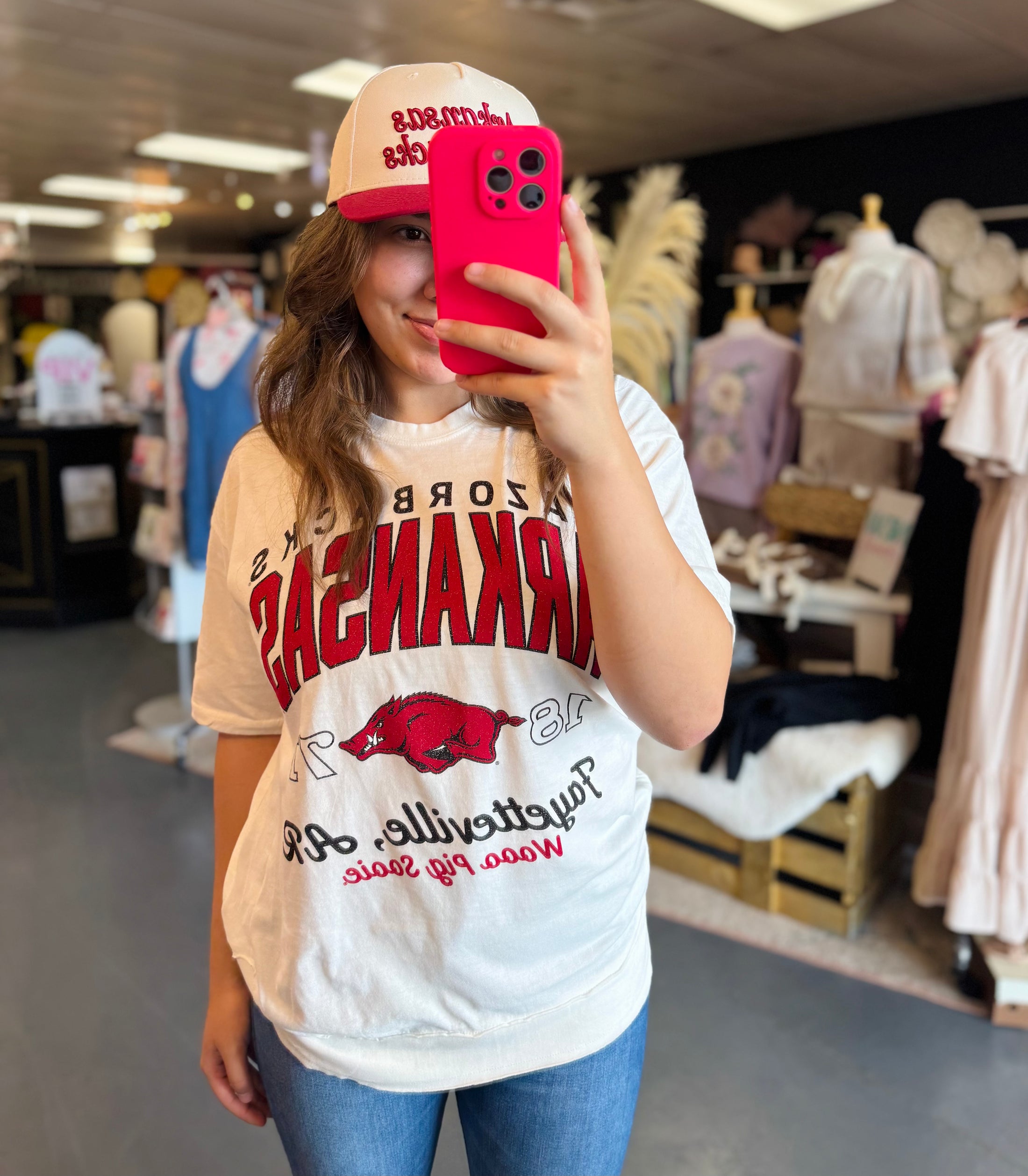Woman wearing a white t-shirt with razorback graphics, taking a mirror selfie in a Fayetteville store.