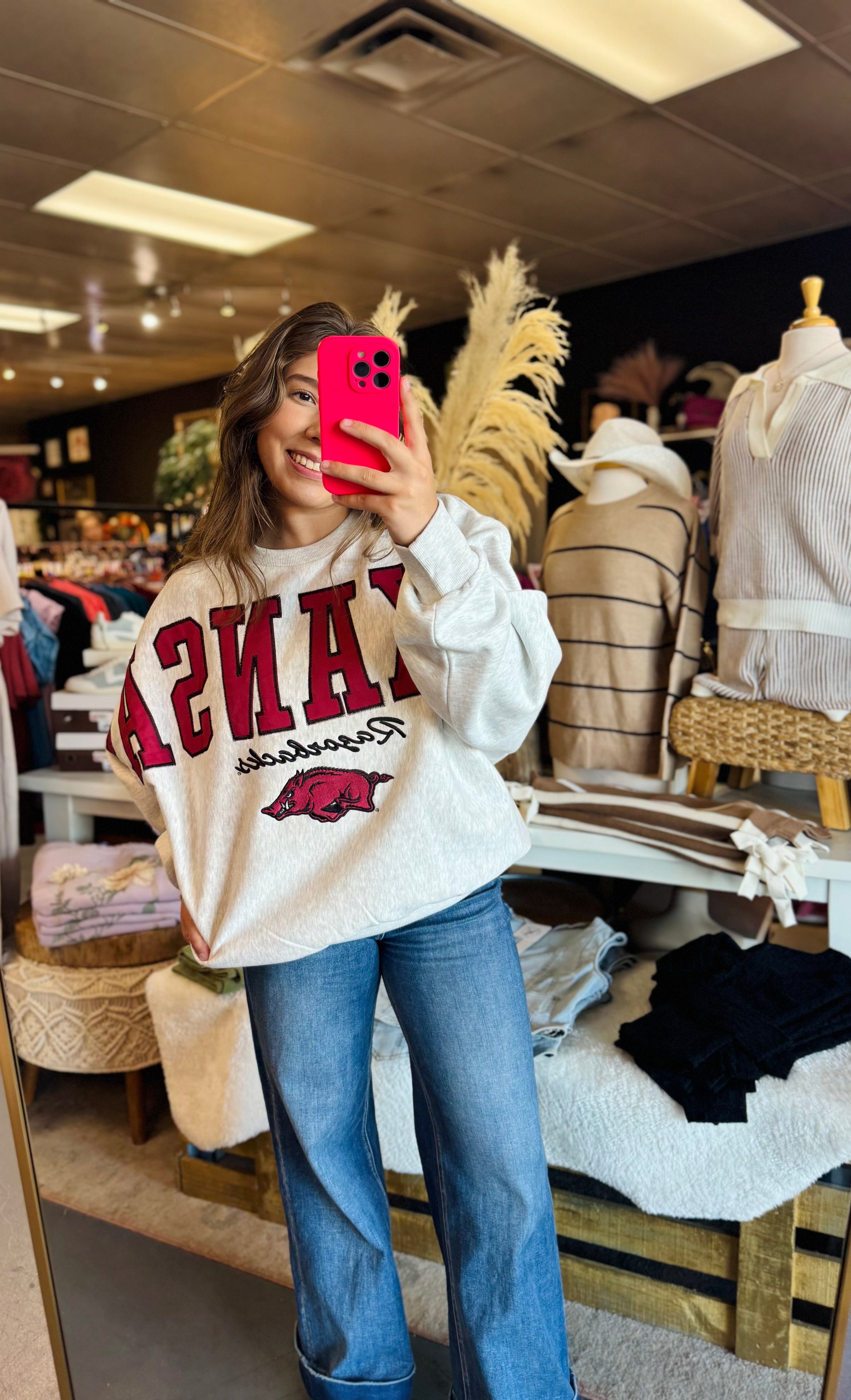 Women wearing a white arkansas sweatshirt with razorback embroidery and hogs logo, taking a selfie in a Fayetteville arkansas store.  Boutique razorback clothing near me, arkansas razorback store, store near me, Fayetteville ar stores, arkansas razorback store