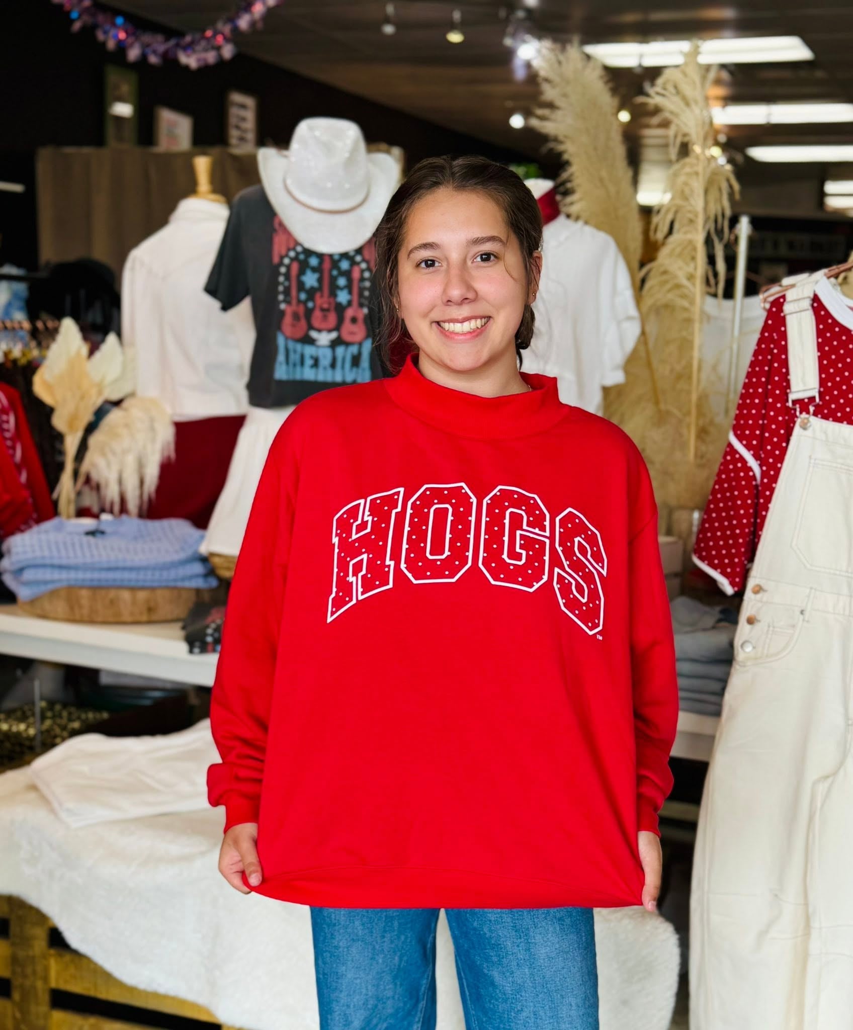 A smiling woman wearing a bright red sweatshirt with 'HOGS' in large white varsity-style letters across the chest, standing in a Fayetteville boutique retail store.