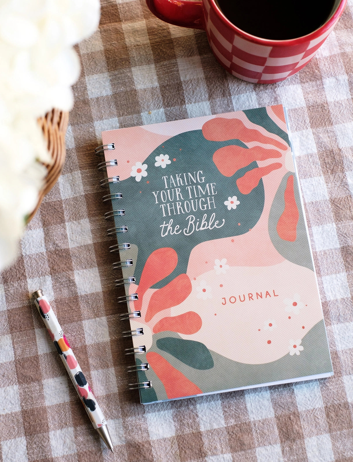 A spiral-bound journal with a multicolored floral design on the cover, titled 'Taking Your Time Through the Bible Journal', displayed on a table with a cup of coffee and a pen. Taking your time through the Bible journal, bible study journal, journey journal, christian journal