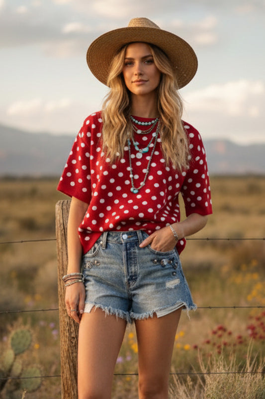 A woman modeling the "My Hog Calling Polka Dot Sweater" tucked into distressed denim shorts and accessorized with layered turquoise necklaces and a straw hat. An essential piece for the Whimsy Boutique "Red Edit" collection.