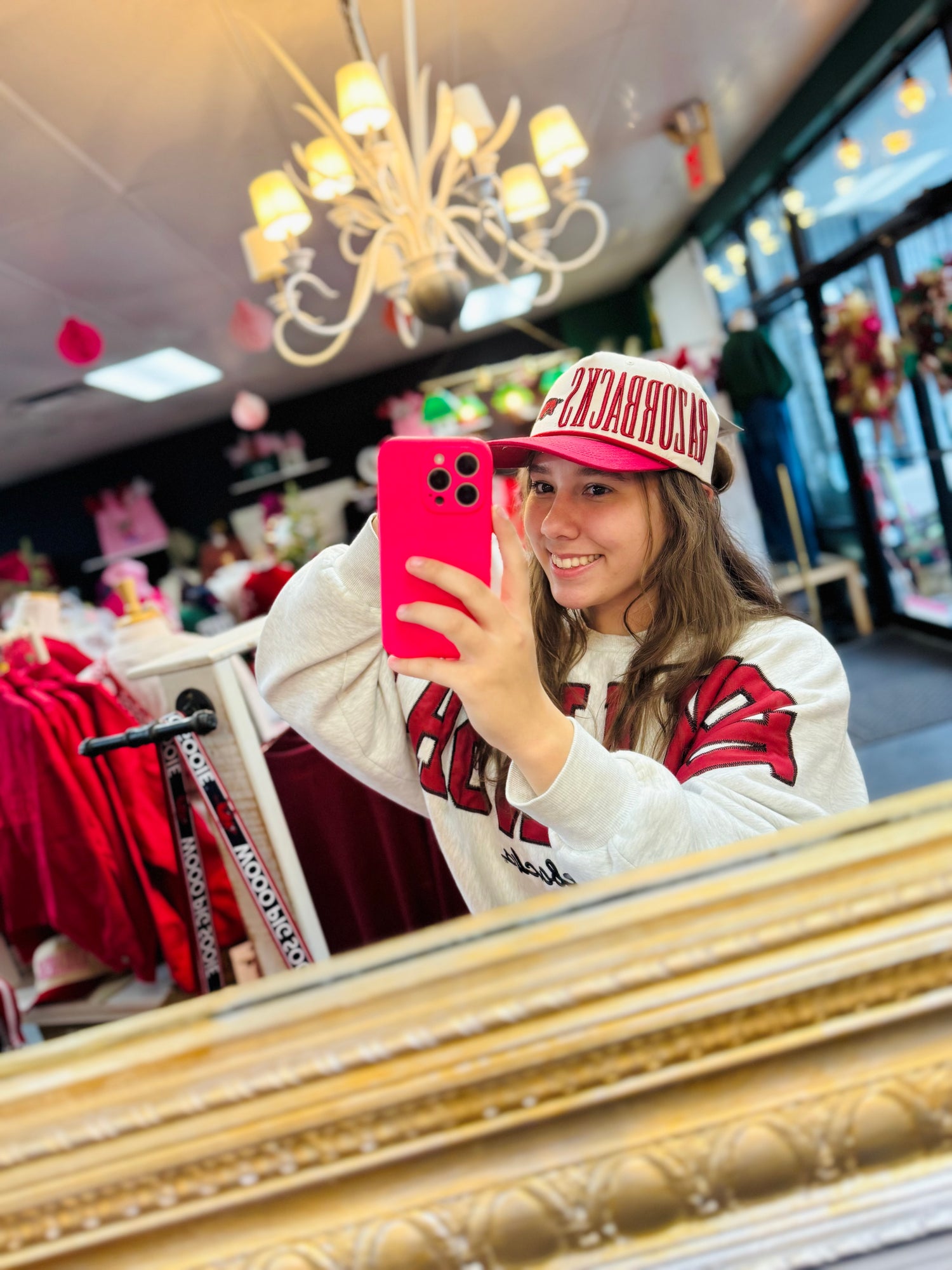 Young woman taking a mirror selfie wearing a red and white Razorback trucker hat and a vintage-style Arkansas sweatshirt at Whimsy Whoo in Fayetteville.