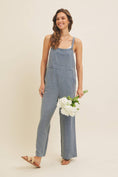 Load image into Gallery viewer, Woman wearing a blue linen-blend jumpsuit holding white flowers against a beige background

