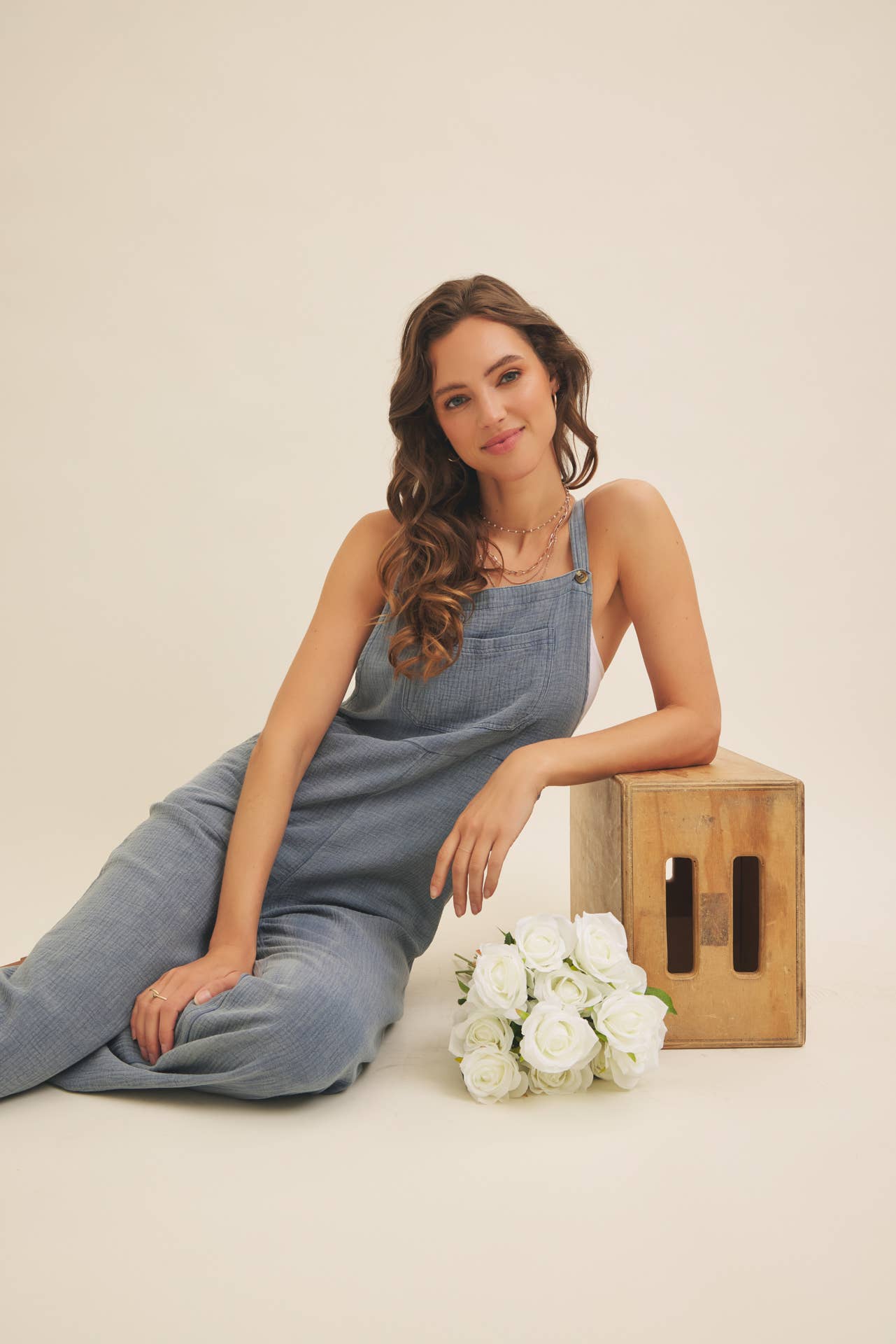 Woman in a denim jumpsuit sitting on a wooden box with flowers, against a plain background